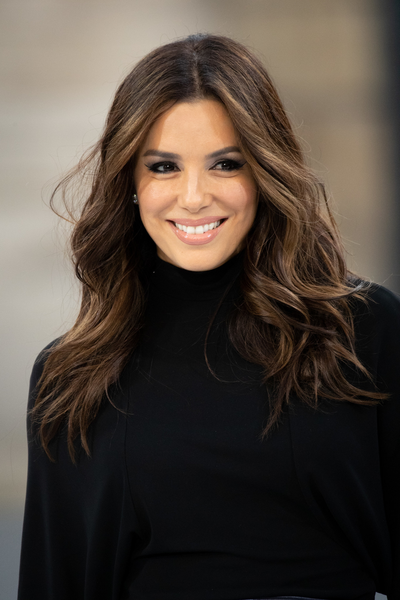 Smiling woman with long wavy brown hair wearing a black top against a blurred background.