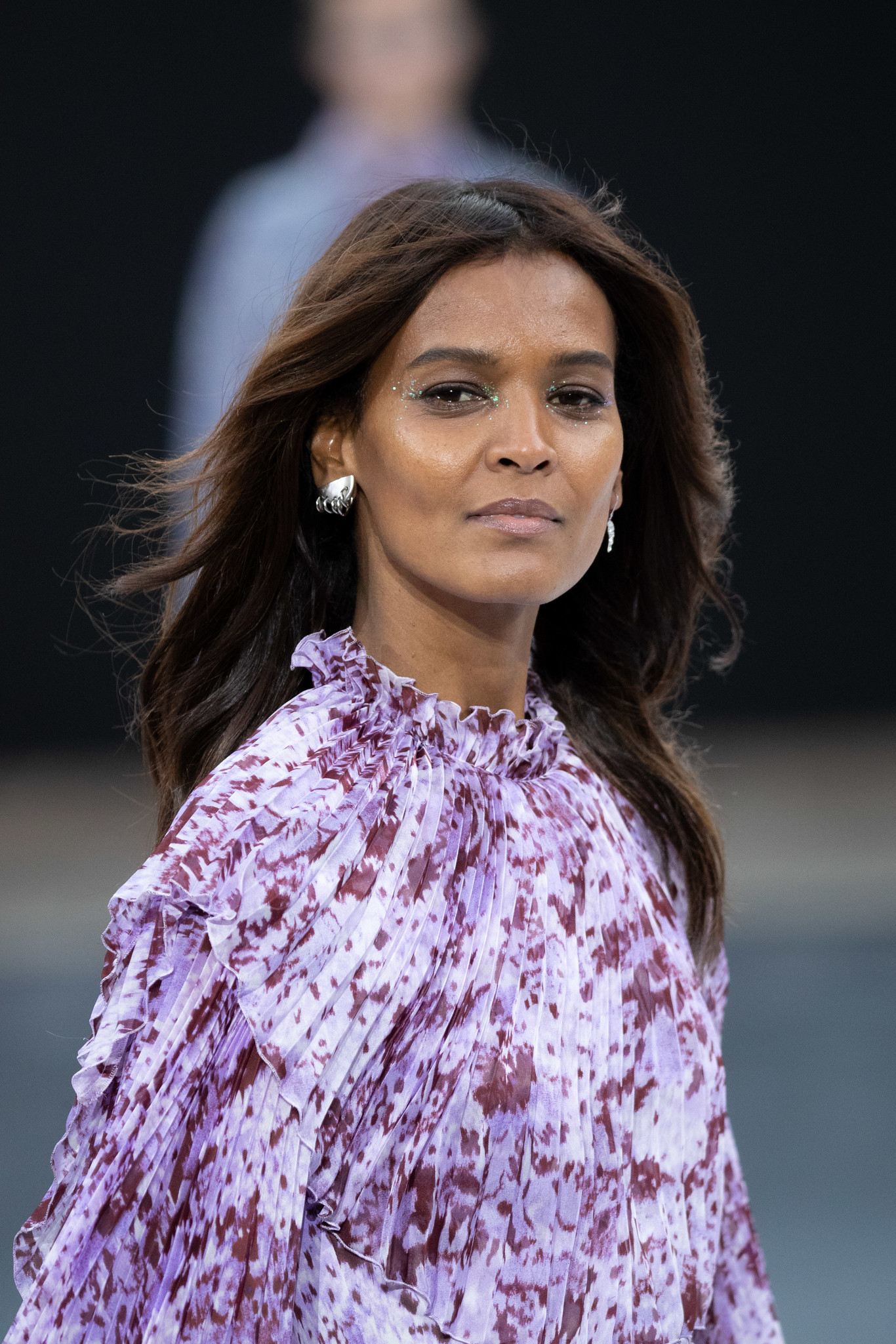 Woman with long brown hair wearing a ruffled purple and white patterned blouse with subtle glitter makeup near her eyes.
