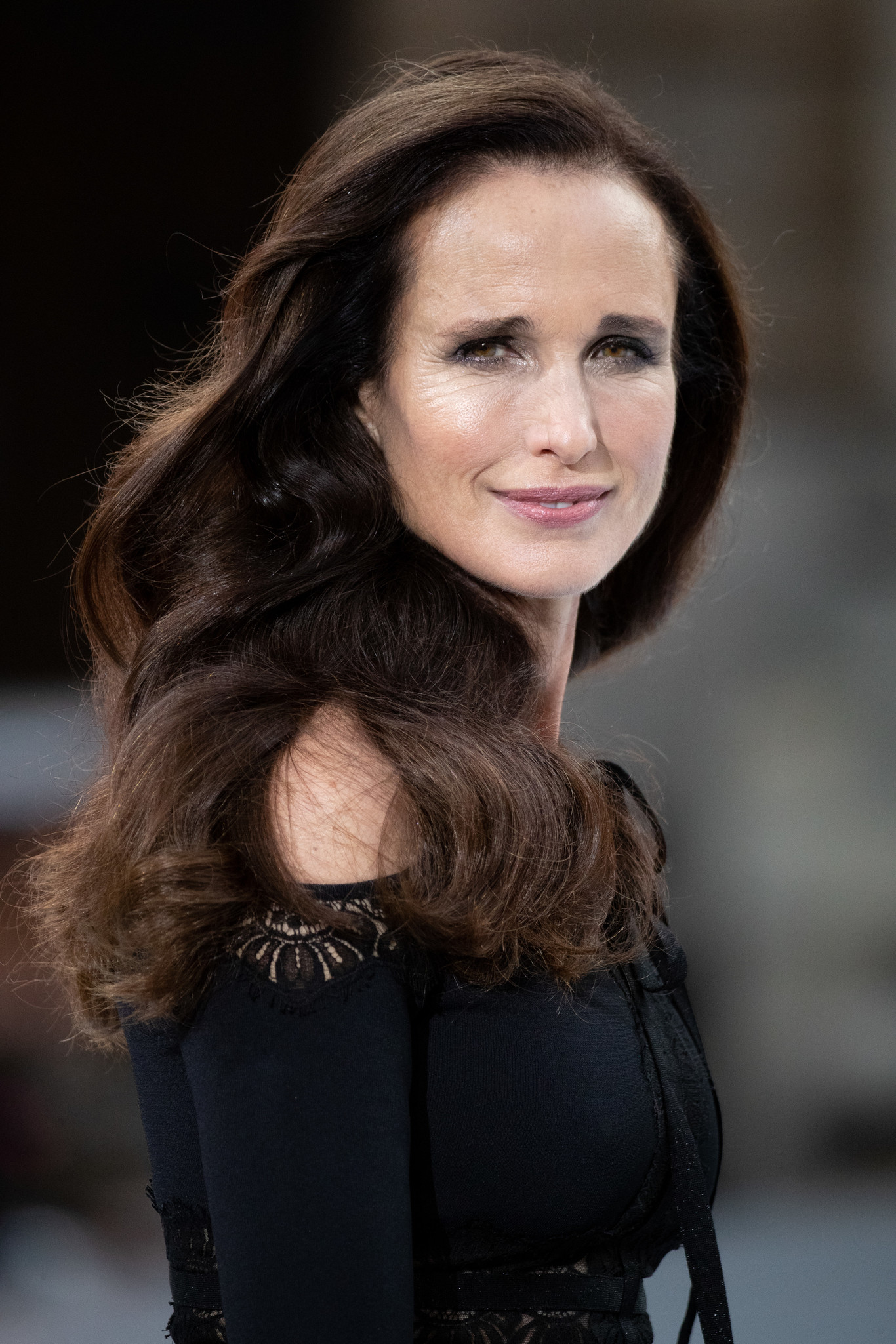 Woman with long wavy brown hair wearing a black off-shoulder dress with lace details, smiling slightly and looking at the camera.